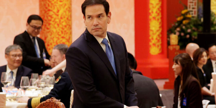 Marco Rubio in a dark suit stands beside an ornately carved chair at a formal event with red and gold decor. Several people are seated at tables in the background, dressed in formal attire.