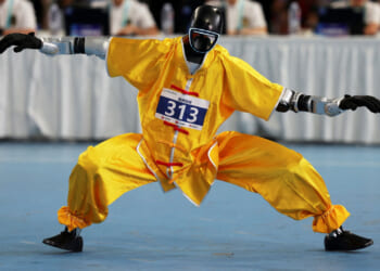 A humanoid robot in yellow martial arts attire and protective gear strikes a low, wide stance on a blue floor during a competition in China, with judges seated at a table in the background.