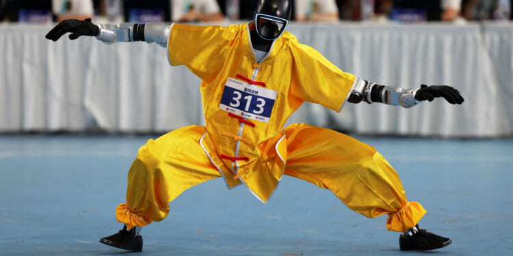 A humanoid robot in yellow martial arts attire and protective gear strikes a low, wide stance on a blue floor during a competition in China, with judges seated at a table in the background.