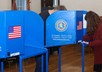 Two people fill out ballots at blue voting booths in a room with wood floors and walls. The booths display the Nassau County seal, an American flag sticker, and a sign with voter information in English and Spanish.