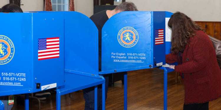 Two people fill out ballots at blue voting booths in a room with wood floors and walls. The booths display the Nassau County seal, an American flag sticker, and a sign with voter information in English and Spanish.