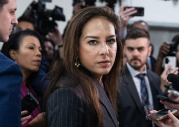 Justice Department Civil Rights Division head Harmeet Dhillon announced a lawsuit over transgender policies in Kansas. Pictured: Dhillon, a woman in a dark suit stands in a crowded room, surrounded by reporters holding microphones and cameras, looking directly at the camera with a serious expression.