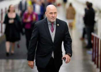 Rep. Clay Higgins in a suit and red tie walks indoors, holding a phone. People are blurred in the background, and the setting appears to be a hallway or corridor with white walls and a shiny floor.