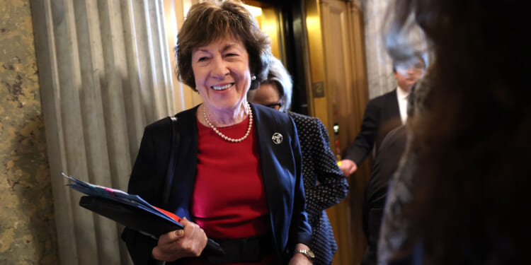 Sen. Susan Collins in a red dress and black blazer, smiling, holds folders while standing near an elevator in a building with marble columns. Other people are around her.