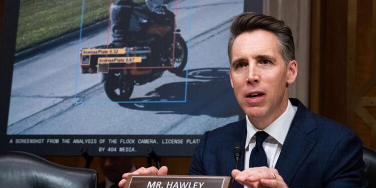 A man in a suit speaks at a hearing, seated at a desk labeled Mr. Hawley. Behind him, a screen displays a vehicle image with highlighted license plate data and analysis from a camera system.