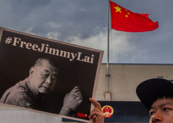 A person holds a #FreeJimmyLai sign with a black-and-white photo of a man. In the background, a Chinese flag flies above a building under a cloudy sky.