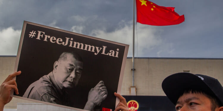 A person holds a #FreeJimmyLai sign with a black-and-white photo of a man. In the background, a Chinese flag flies above a building under a cloudy sky.