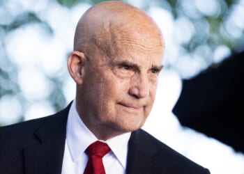 A bald man in a suit and red tie stands outdoors, looking to the side with a slight smile. The background is blurred with green and white bokeh from trees.