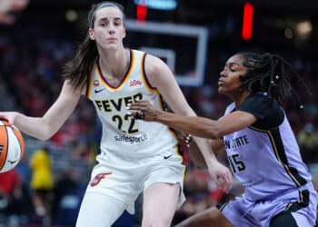 Caitlin Clark in a white Fever uniform dribbles while being closely guarded by a defender in a purple uniform during a game, with a blurred crowd in the background.
