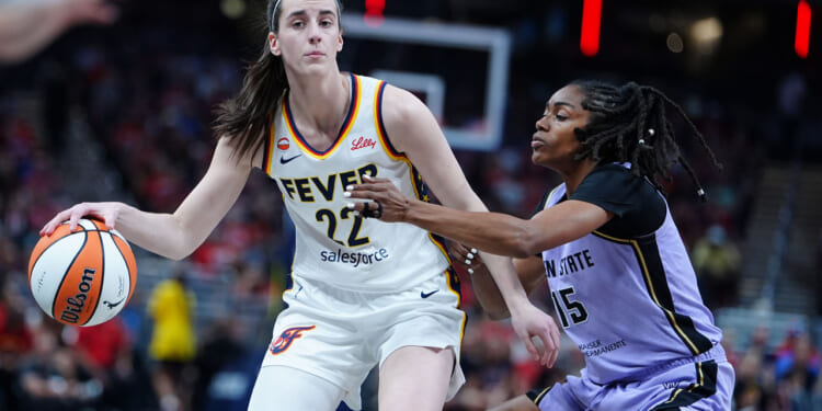 Caitlin Clark in a white Fever uniform dribbles while being closely guarded by a defender in a purple uniform during a game, with a blurred crowd in the background.