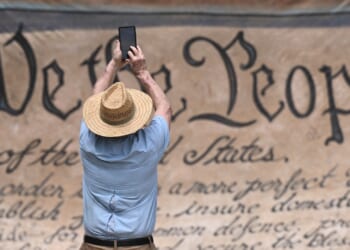 A person in a straw hat takes a photo with a phone in front of a large replica of the U.S. Constitutions We the People preamble.