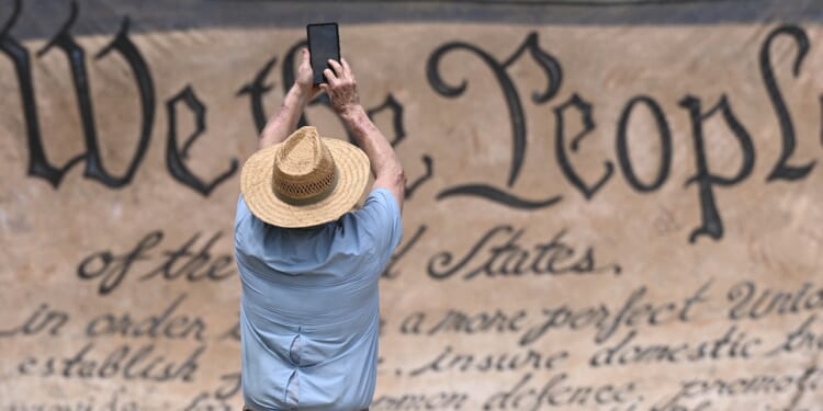 A person in a straw hat takes a photo with a phone in front of a large replica of the U.S. Constitutions We the People preamble.