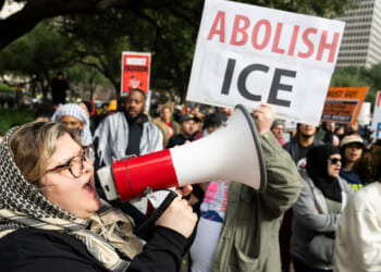 A woman shouts into a megaphone at a protest. Behind her, people hold signs, including one that says ABOLISH ICE in red and white. The crowd appears passionate and engaged. Education News