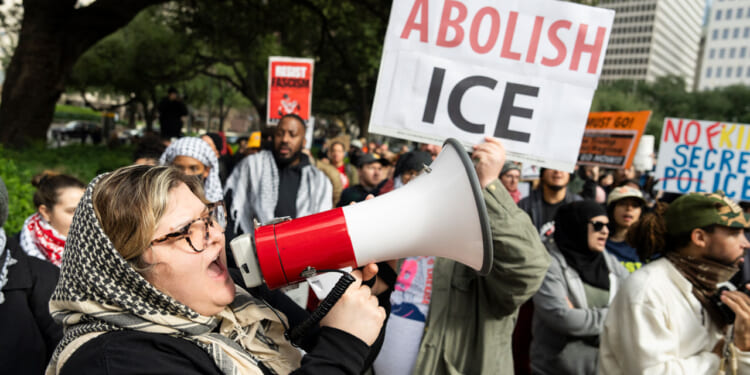 A woman shouts into a megaphone at a protest. Behind her, people hold signs, including one that says ABOLISH ICE in red and white. The crowd appears passionate and engaged. Education News