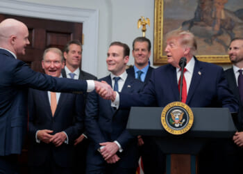 A group of men in suits, including Donald Trump at a podium, smile as two shake hands in front. The podium with the U.S. presidential seal hints at an important announcement—as a large painting hangs on the wall behind them.