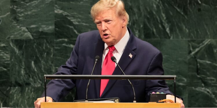 A man in a suit and red tie speaks at a podium in front of a green marble wall, appearing to address an assembly or conference.