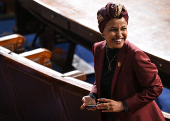 Ilhan Omar in a maroon blazer and patterned headwrap smiles while standing in a large, formal room with wooden seats and a round table, holding a phone.
