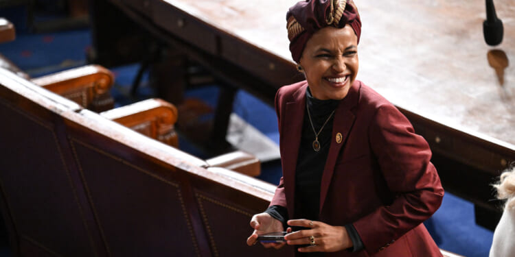 Ilhan Omar in a maroon blazer and patterned headwrap smiles while standing in a large, formal room with wooden seats and a round table, holding a phone.
