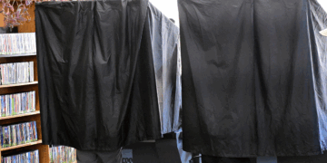 Two people stand inside separate curtained voting booths. The black curtains provide privacy, and bookshelves filled with DVDs are visible in the background.