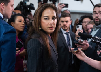 Harmeet Dhillon in a dark suit stands surrounded by reporters holding microphones and recording devices, while cameras and journalists focus on her in a crowded hallway.