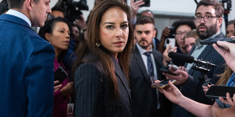 Harmeet Dhillon in a dark suit stands surrounded by reporters holding microphones and recording devices, while cameras and journalists focus on her in a crowded hallway.