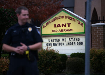A police officer stands in front of a sign for the Islamic Association of North Texas that reads, United We Stand, One Nation Under God.