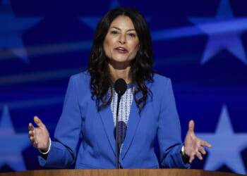 Michigan Attorney General Dana Nessel. Pictured: a woman in a blue blazer speaks at a podium with a microphone, gesturing with her hands. The background displays large white stars on a blue backdrop.
