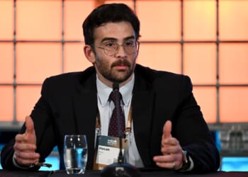 A man in a suit and glasses speaks at a conference table with a microphone, notepad, and a glass of water in front of him. An orange and blue grid background is visible behind him.