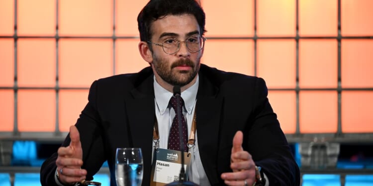 A man in a suit and glasses speaks at a conference table with a microphone, notepad, and a glass of water in front of him. An orange and blue grid background is visible behind him.