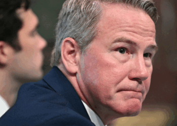 A man with short gray hair in a blue suit looks slightly to the side with a serious expression. Another person sits blurred in the background. An American flag is partially visible on the right.