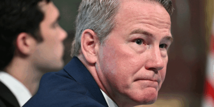 A man with short gray hair in a blue suit looks slightly to the side with a serious expression. Another person sits blurred in the background. An American flag is partially visible on the right.