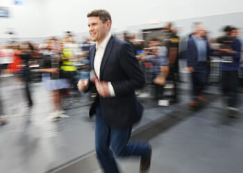 A man in a dark blazer and jeans jogs indoors with a blurred background of people and cameras, giving a sense of motion and energy.
