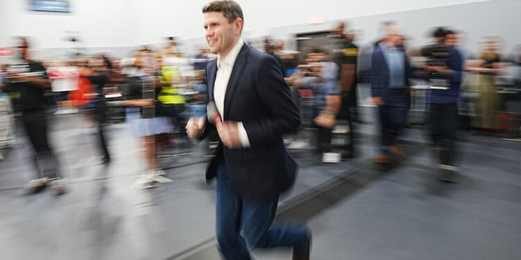 A man in a dark blazer and jeans jogs indoors with a blurred background of people and cameras, giving a sense of motion and energy.