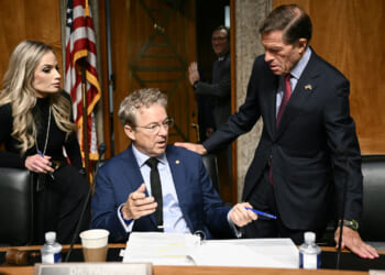 Three people in a formal meeting room talk around a desk with papers, coffee cups, and pens; an American flag is in the background.
