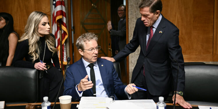 Three people in a formal meeting room talk around a desk with papers, coffee cups, and pens; an American flag is in the background.