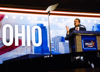 A person in a suit speaks at a podium labeled Dallas, with OHIO and a city skyline in the background. A teleprompter is visible overhead.