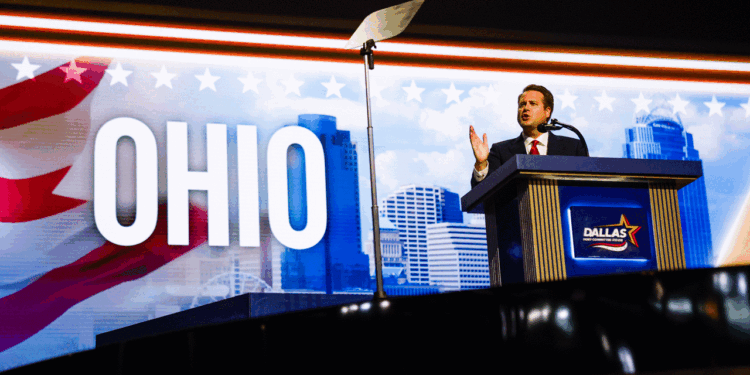 A person in a suit speaks at a podium labeled Dallas, with OHIO and a city skyline in the background. A teleprompter is visible overhead.