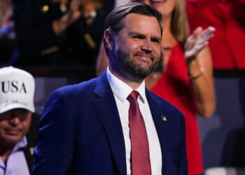 A man in a blue suit and red tie smiles while standing at an event. People are visible in the blurred background, including someone in a white USA hat.