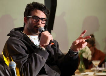 A piker wearing glasses and a dark jacket speaks into a microphone, gesturing with two fingers. There are drinks and flowers on the table in the background, and another person is faintly visible.