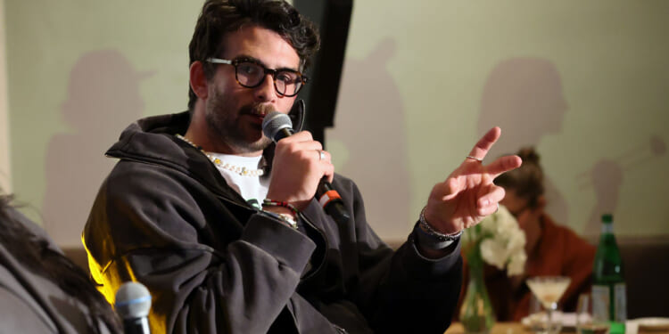 A piker wearing glasses and a dark jacket speaks into a microphone, gesturing with two fingers. There are drinks and flowers on the table in the background, and another person is faintly visible.