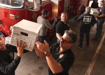 Firefighters form a human chain, passing white boxes in front of a red fire truck inside a fire station. Some wear dark shirts with FIRE on the back; one wears sunglasses.