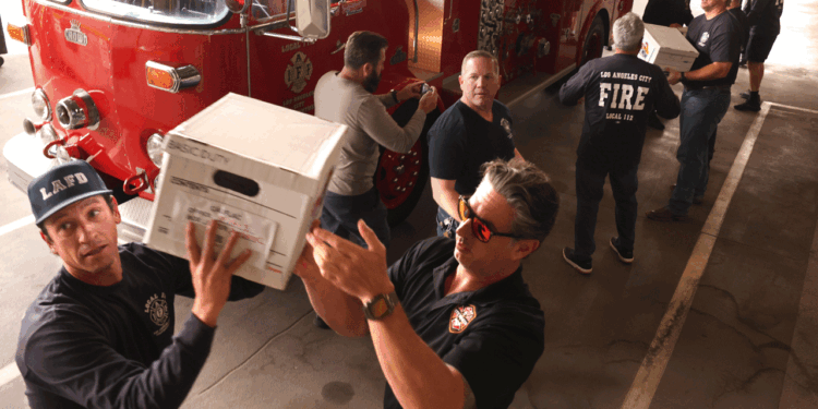Firefighters form a human chain, passing white boxes in front of a red fire truck inside a fire station. Some wear dark shirts with FIRE on the back; one wears sunglasses.