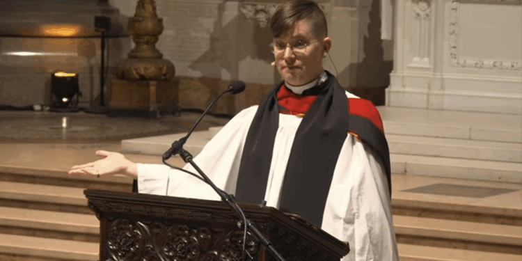 Rev. Flouish Klink claims Jesus is trans. Pictured: a person in religious robes stands at a carved wooden pulpit, speaking into a microphone, with one hand gesturing outward inside a large, ornate church.
