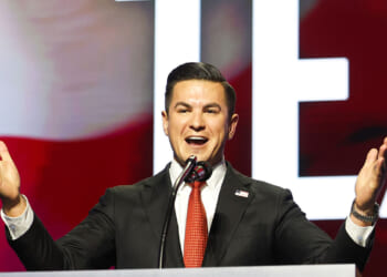 A man in a suit speaks enthusiastically at a podium with a microphone, gesturing with open arms. The background displays large, bold letters and red, white, and blue colors.