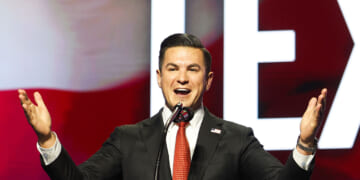 A man in a suit speaks enthusiastically at a podium with a microphone, gesturing with open arms. The background displays large, bold letters and red, white, and blue colors.