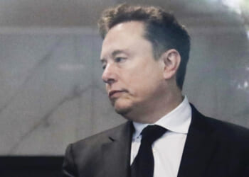 Elon Musk in a black suit and tie looks to the side with a neutral expression while standing indoors.