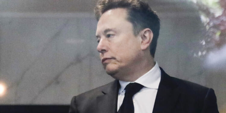 Elon Musk in a black suit and tie looks to the side with a neutral expression while standing indoors.
