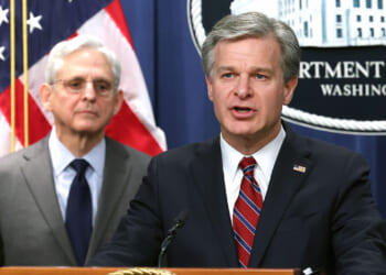 FBI Director Chris Wray and Merrick Garland. Two men in suits stand at a podium. One is speaking, the other stands behind him. An American flag and a Department of Justice sign are visible in the background.