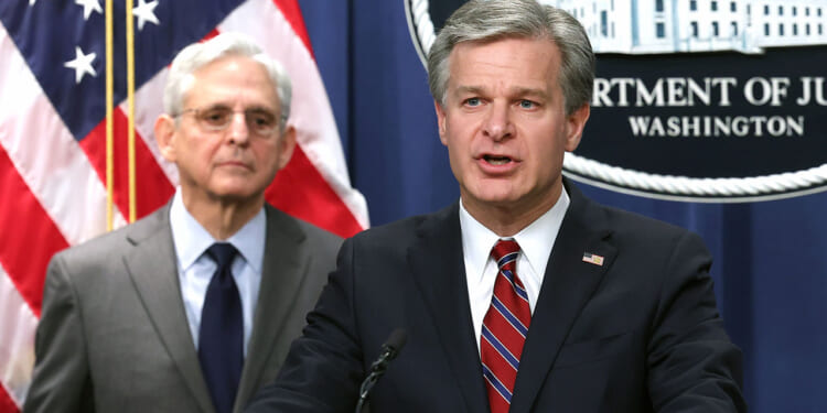 FBI Director Chris Wray and Merrick Garland. Two men in suits stand at a podium. One is speaking, the other stands behind him. An American flag and a Department of Justice sign are visible in the background.