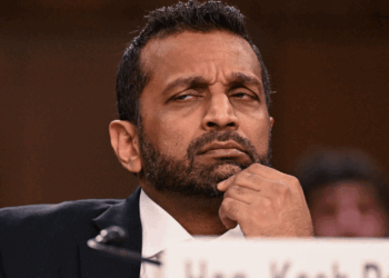 Kash Patel in a suit sits with his hand on his chin, looking skeptical or thoughtful during a formal event. A blurred background and a partial nameplate are visible in the foreground.